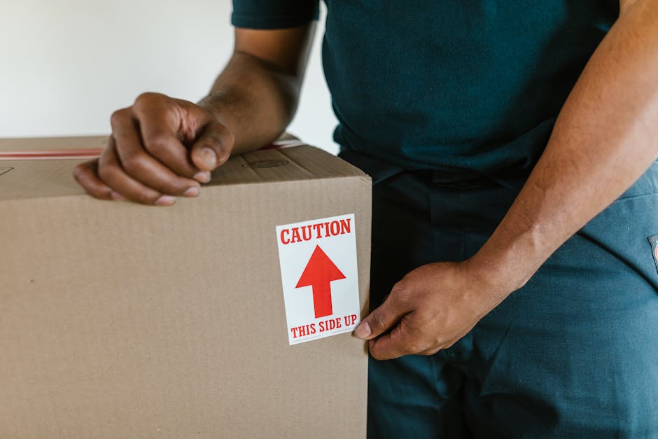 A man standing indoors among numerous brown cardboard boxes of various sizes, some sealed with red packing tape and others partially open, revealing packaging materials. He is holding a smartphone in