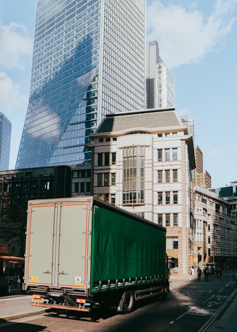 A large green moving truck with a closed cargo area is parked on a city street in front of modern skyscrapers and historic office buildings. The truck is positioned near the curb, with the rear doors